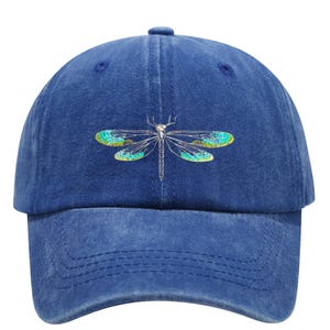 May include: A blue baseball cap with a dragonfly design. The dragonfly has teal and yellow wings and a silver body. The cap is made of a soft, textured material and has a curved brim.