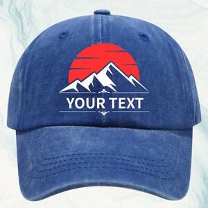 Puede incluir: Gorra de béisbol azul con un gráfico de montaña y sol. El gráfico presenta un sol rojo, montañas blancas y el texto "YOUR TEXT" en blanco. La gorra está hecha de un material suave y tiene una visera curva.