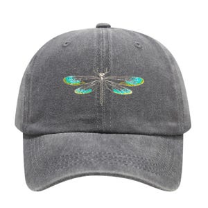 May include: A gray baseball cap featuring an embroidered dragonfly design. The dragonfly has teal and yellow wings and a silver body. The cap has a curved brim and a fabric strap closure.