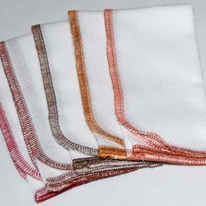 Puede incluir: Cinco paños de cocina blancos con bordes de diferentes colores. Los bordes son rojos, marrones, naranjas y naranjas claros. Los paños están doblados por la mitad y apilados uno encima del otro.