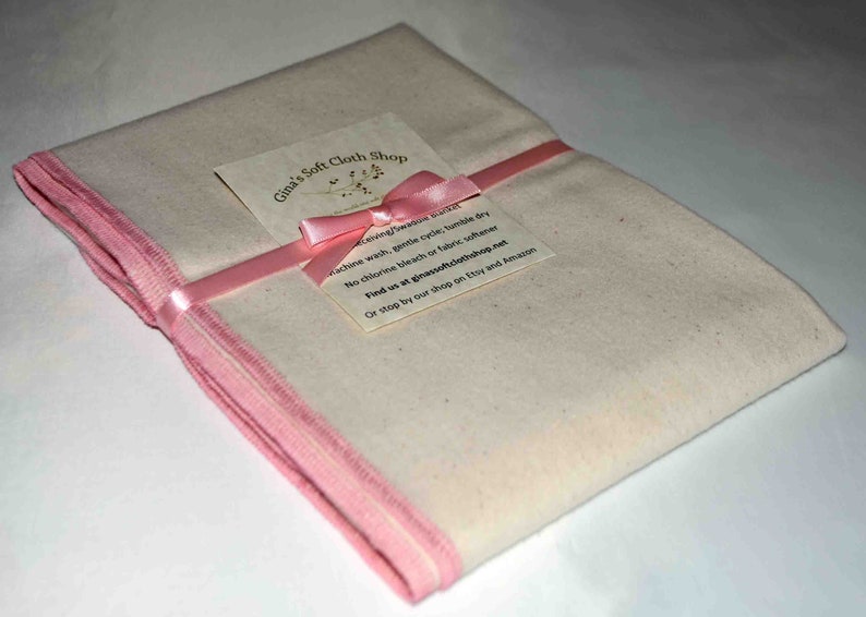 May include: A folded white cloth with pink trim, tied with a pink ribbon and a pink bow. A small card with the text "Gina's Soft Cloth Shop" is attached to the ribbon. The card includes washing instructions: "Machine wash, gentle cycle, tumble dry. No chlorine bleach or fabric softener. Find us at ginasoftclothshop.net. Or stop by our shop on Etsy and Amazon."