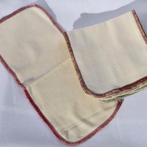May include: Two sets of white cotton cloths with red, green, and yellow stitching around the edges. The cloths are folded and stacked on top of each other.