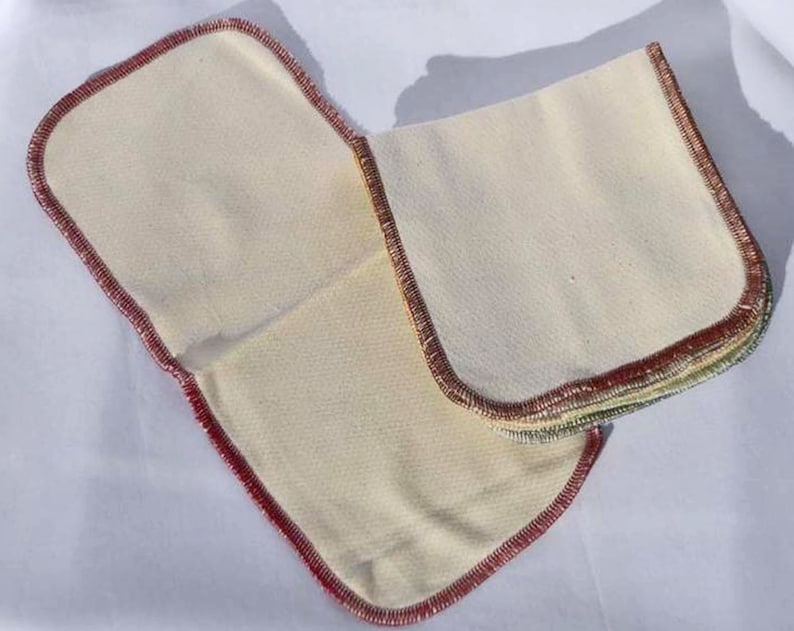 May include: Two sets of white cotton cloths with red, green, and yellow stitching around the edges. The cloths are folded and stacked on top of each other.