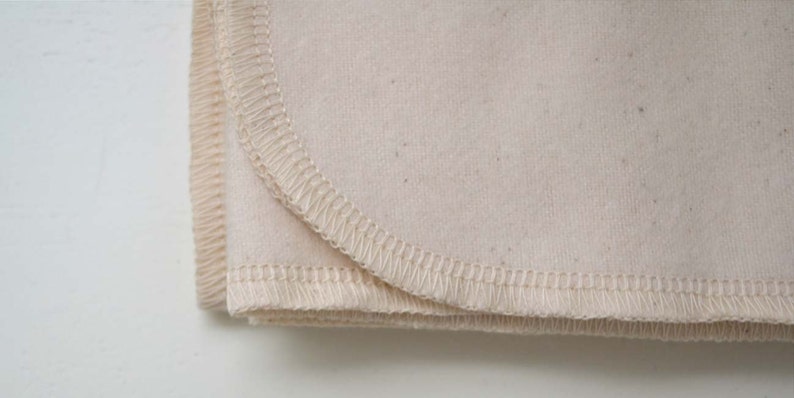 May include: Close-up of a folded piece of off-white fabric with a stitched edge. The fabric is soft and has a slightly textured surface.