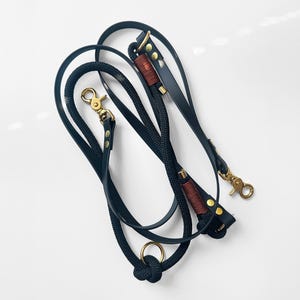 Op de afbeelding: Een zwarte hondenriem met een touwontwerp, leren accenten en goudkleurige metalen hardware. De riem heeft een knoop aan de basis en een sluiting aan de bovenkant. De leren accenten zijn bruin. Voor dierenbezitters.