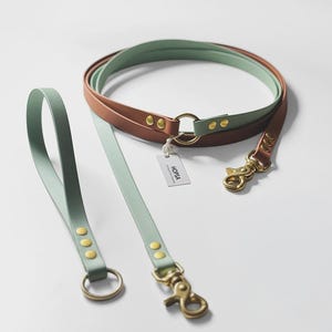 Könnte beinhalten: Eine zweifarbige Hundeleine mit einem mintgrünen und terrakottabraunen Riemen. Die Leine verfügt über messingfarbene Metallbeschläge, darunter drehbare Karabinerhaken, Nieten und Ringe. Ein kleines weißes Etikett mit dem Text HOPSA ist befestigt.