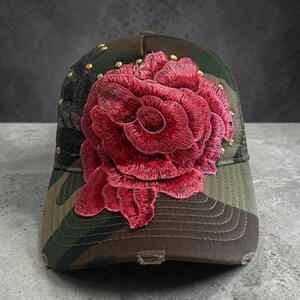 May include: A camouflage baseball cap featuring a large, vibrant red embroidered rose appliqué on the front panel. The cap includes small gold-toned metal studs scattered around the floral design and shows distressed detailing along the brim edge.