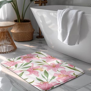 Könnte beinhalten: Eine weiße Badematte mit einem rosafarbenen Blumenmuster mit Lilien. Die Matte ist aus weichem, saugfähigem Material gefertigt und perfekt, um jedem Badezimmer einen Hauch von Eleganz zu verleihen.