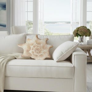 Pode incluir: Um sofá creme com várias almofadas e uma almofada decorativa em forma de concha. Uma manta bege está sobre o sofá. O quarto tem um tema costeiro com uma grande janela com vista para a água.