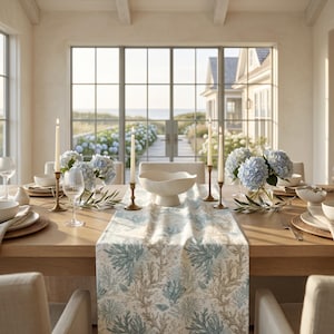 May include: A wooden dining table set with a table runner featuring a blue and beige coral pattern. The table includes a white pedestal bowl, four tall taper candles in brass holders, clear ribbed glassware, white plates, and blue hydrangea bouquets.