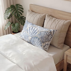May include: A bed with a light wood headboard and a white duvet. Two beige quilted pillows are against the headboard. A rectangular pillow with a blue and white seashell pattern is on top of the duvet. A potted plant is in the background.