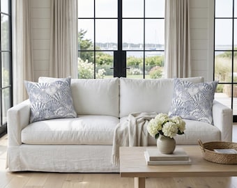 May include: A white sectional sofa with two decorative pillows featuring a blue and white seashell pattern. A beige throw blanket rests on the sofa. A wooden coffee table holds a vase of white flowers and a woven basket.