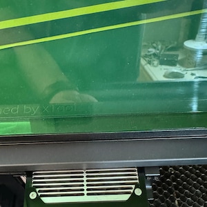 May include: A close-up of a black laser engraver with a 20W laser power output. The laser engraver has a metal grate and a green protective cover.