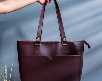 May include: A burgundy leather tote bag with two handles and a rectangular shape. The bag has a front pocket and a zipper closure. The handles are attached with gold-colored hardware. The bag is held up by a hand with red nail polish.