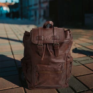 May include: A brown leather backpack with a flap closure and buckle straps. The backpack has a top handle, side pockets, and a front zippered pocket. The bag has the letters "AD" embossed on the flap.