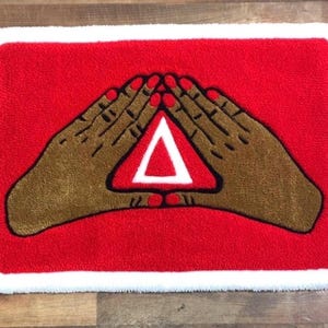 Puede incluir: Una alfombra rectangular roja con borde blanco. Dos manos marrones con esmalte de u&ntilde;as rojo forman un tri&aacute;ngulo alrededor de un s&iacute;mbolo delta blanco. La alfombra tiene una apariencia texturizada.