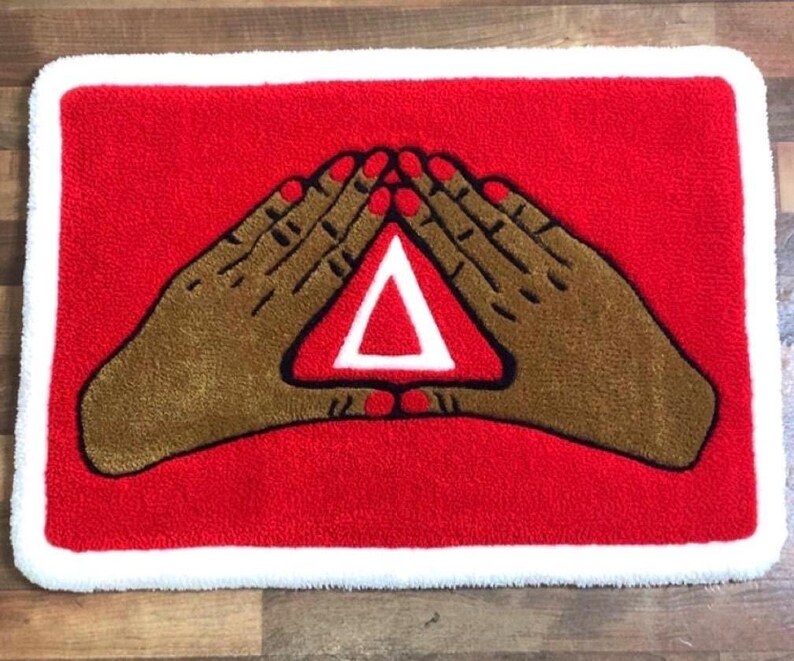 Puede incluir: Una alfombra rectangular roja con borde blanco. Dos manos marrones con esmalte de u&ntilde;as rojo forman un tri&aacute;ngulo alrededor de un s&iacute;mbolo delta blanco. La alfombra tiene una apariencia texturizada.