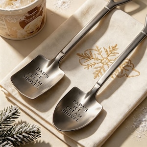 May include: Two matte silver metal spoons shaped like miniature shovels with triangular handles. One spoon is engraved with the text DAD'S ICE CREAM SHOVEL and the other is engraved with the text PAPA'S ICE CREAM SHOVEL, both resting on a white cloth.