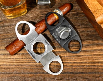 May include: Two cigar cutters, one black and one silver, with the word "Groomsmen" engraved on each. The black cutter has the word "Philips" engraved on it. The silver cutter has the word "Philips" engraved on it. A brown cigar is lying on a wooden surface.