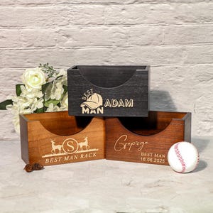Puede incluir: Tres organizadores de escritorio de madera con aberturas frontales curvas. Uno es negro con el texto "BEST MAN ADAM", otro es marrón con "BEST MAN RACK" y un diseño de ciervo, y el tercero es marrón con el texto "George BEST MAN 16.06.2025". Una pelota de béisbol está al lado del tercer organizador.