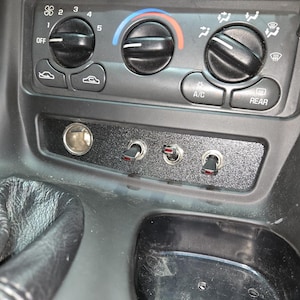 May include: Close-up of a car's interior with a black dashboard, a cigarette lighter, and three toggle switches. The dashboard has a black and silver design with a textured surface. The switches are black with silver trim and are labelled "OFF", "A/C", and "REAR".