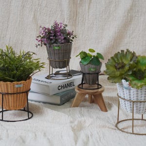 May include: A collection of decorative woven planters with metal stands, each holding different types of artificial plants. The planters are in various colors, including gray, white, and orange, and are arranged on a neutral-toned surface.