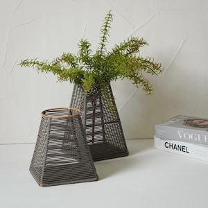 May include: Two gray woven vases, one holding green leafy branches, sit on a white surface. The vases have a geometric, trapezoid shape. A stack of books with the titles "Vogue" and "Chanel" is visible in the background.