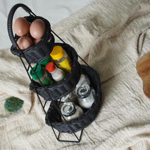 May include: A three-tiered black wicker basket stand filled with eggs, spices, and jars. The top basket holds brown eggs, the middle basket contains spice bottles, and the bottom basket has jars with silver lids. The stand has a handle.