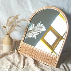 以下が含まれることがあります： アーチ型の装飾ミラー。ベースとフレームは織り込まれたラタン製。鏡は黄色と白の光が差し込む窓を映しています。小さなベージュの花瓶にドライフラワーが活けられており、ミラーの左側に配置されています。