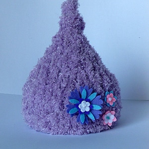 Puede incluir: Un gorro de punto afelpado de color lavanda con la parte superior puntiaguda. El gorro está decorado con tres flores de fieltro en tonos de azul, rosa y blanco. El gorro es probablemente para un bebé o un niño.