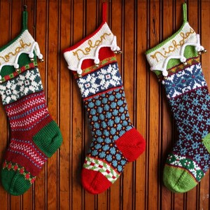 Puede incluir: Tres calcetines de Navidad tejidos cuelgan sobre un fondo de madera. Cada calcetín está personalizado con un nombre en letras doradas. Los calcetines presentan varios patrones en rojo, verde, azul y blanco.