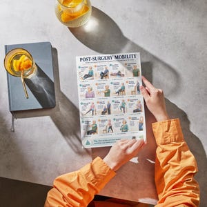 May include: A person holding a printed guide titled "POST-SURGERY MOBILITY" with illustrations of exercises. The guide is on a gray surface, next to a blue notebook and a glass of orange-infused water.