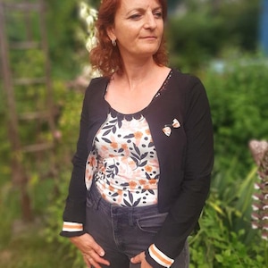 Könnte beinhalten: Eine Frau trägt einen schwarzen Cardigan mit weißen und orangen Ärmelbündchen, ein Oberteil mit Blumendruck und eine dunkelblaue Jeans. Sie trägt eine kleine Schleifenbrosche auf der Vorderseite ihres Cardigans.