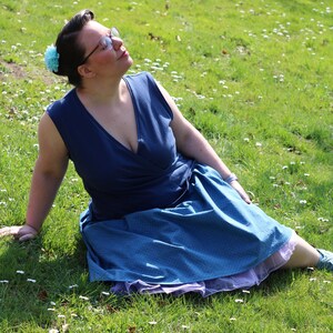 Puede incluir: Una mujer con un vestido azul sin mangas con estampado de lunares blancos se sienta en un campo de hierba. Está mirando hacia el cielo y tiene una flor azul en el pelo.