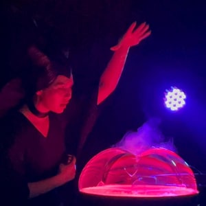 May include: A person wearing a black costume with horns and a red light shining on their face, holding their arm up in the air. A large, clear bubble sits on a black platform with a purple and red light shining through it.