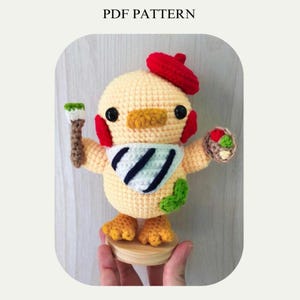 Op de afbeelding: Een gehaakt geel kuiken met een rode baret, een blauw-wit gestreepte sjaal en een penseel en een palet in zijn handen. Het kuiken staat op een houten voet.