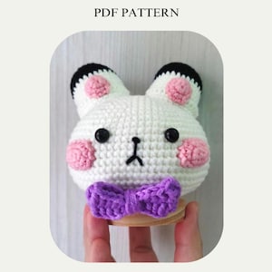 May include: A white crocheted bunny with pink cheeks and black eyes, wearing a purple bow tie. The bunny is sitting on a wooden base.
