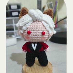 May include: Crocheted doll with a pink face, red eyes, and white hair. The doll wears a black suit with a red trim and a white V-neck. The doll is standing on a wooden base. The text "LOVE AND DEEPSPACE PDF PATTERN" is at the top.