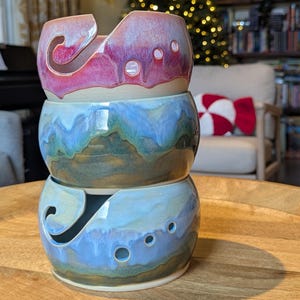 May include: Three glazed ceramic yarn bowls stacked on top of each other. The top bowl is pink and red, the middle bowl is blue and green, and the bottom bowl is blue and tan. Each bowl has a curved slot for yarn and several small holes.