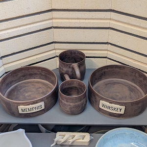 May include: A collection of handmade brown ceramic pottery, including two bowls with the text "MEMPHIS" and "WHISKEY", a mug, and a small cup. The pottery has a rustic, textured appearance. The background features a kiln.