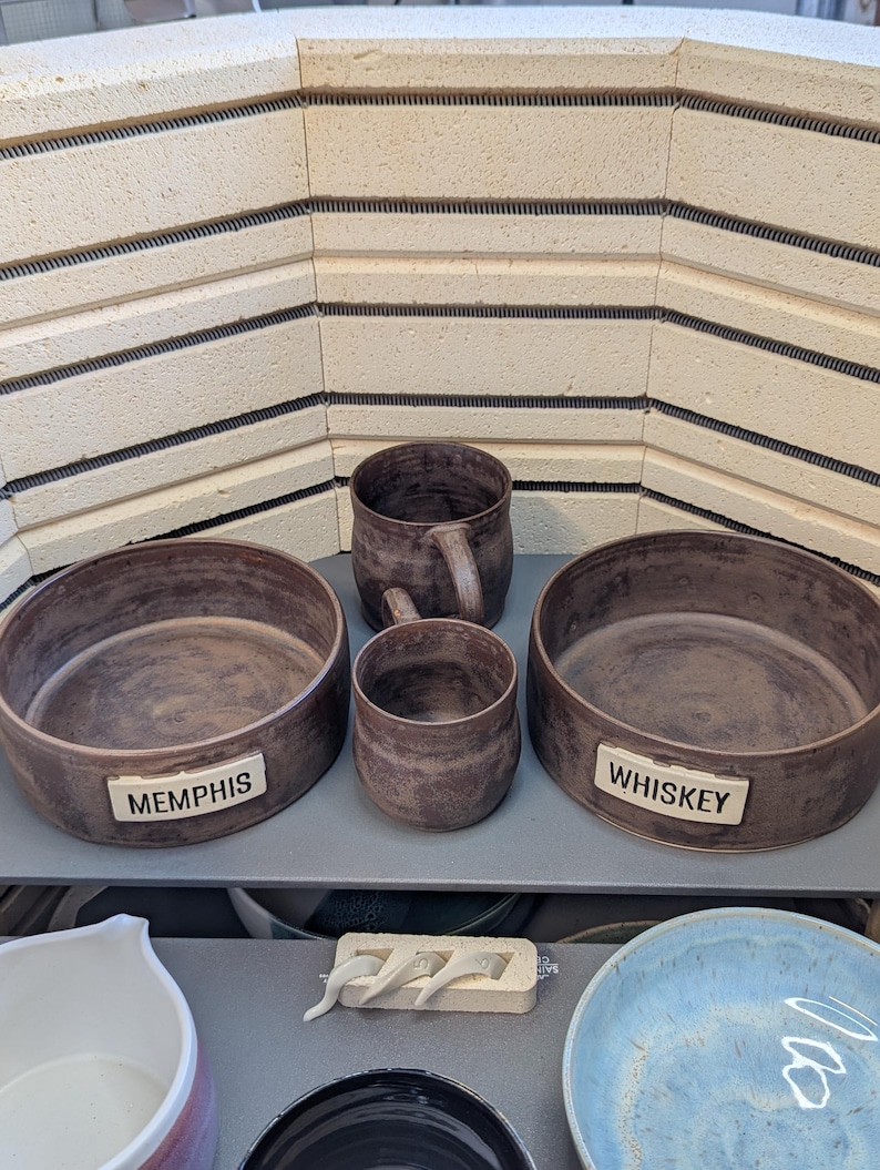 May include: A collection of handmade brown ceramic pottery, including two bowls with the text "MEMPHIS" and "WHISKEY", a mug, and a small cup. The pottery has a rustic, textured appearance. The background features a kiln.