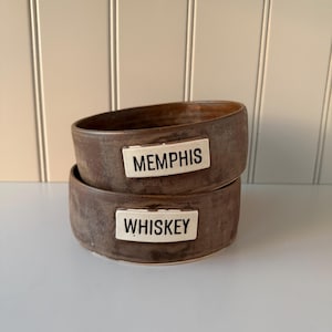 May include: Two stacked, handcrafted brown ceramic bowls. The top bowl has a white label that reads "MEMPHIS." The bottom bowl has a white label that reads "WHISKEY." The bowls have a rustic, earthy aesthetic.