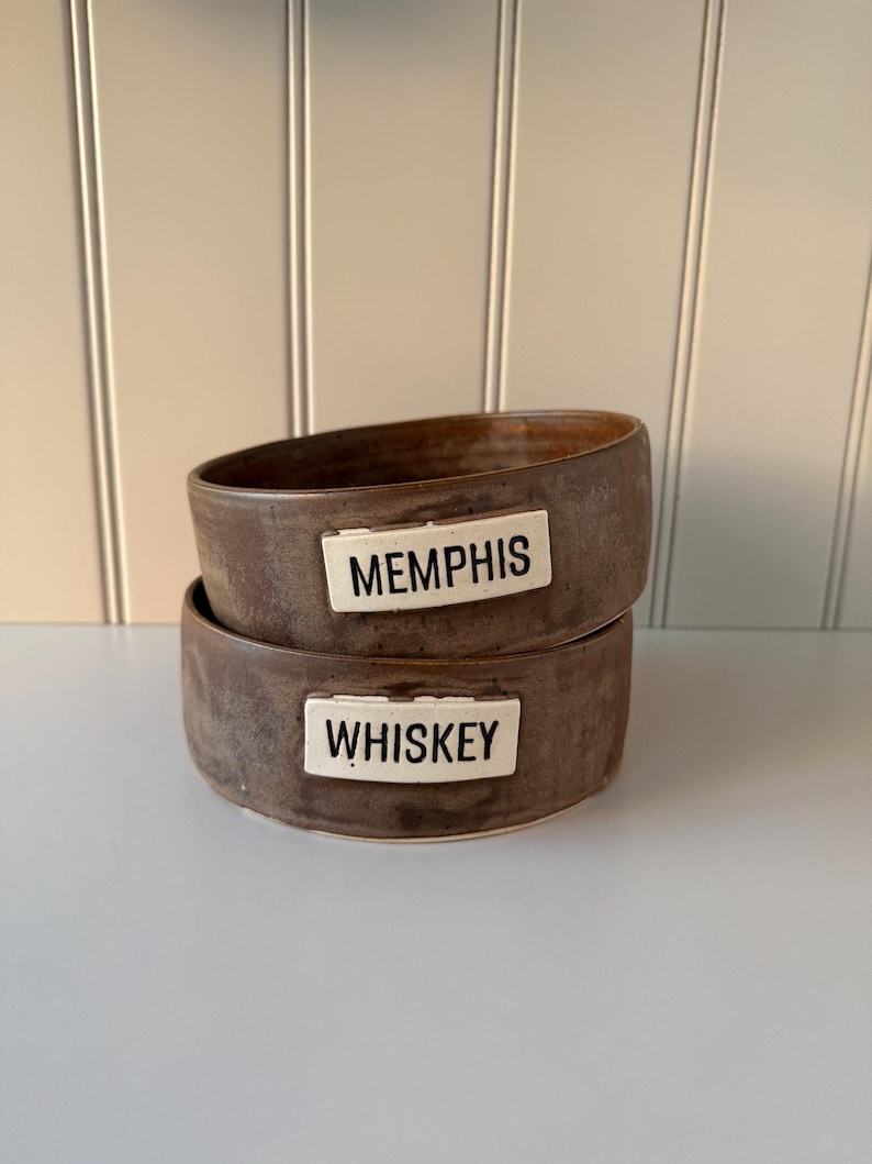 May include: Two stacked, handcrafted brown ceramic bowls. The top bowl has a white label that reads "MEMPHIS." The bottom bowl has a white label that reads "WHISKEY." The bowls have a rustic, earthy aesthetic.