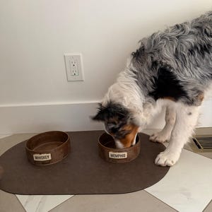 May include: A dog eats from a brown metal bowl labeled "Memphis" on a brown mat. Another bowl labeled "Whiskey" sits nearby. The dog has a mix of black, white, and brown fur. The bowls are on a tiled floor.