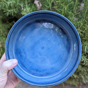 May include: A handmade, round ceramic bowl with a deep blue glaze. The bowl has a slightly textured surface and a wide rim with a lighter blue accent. The bowl is held in a hand, with a green leafy background.