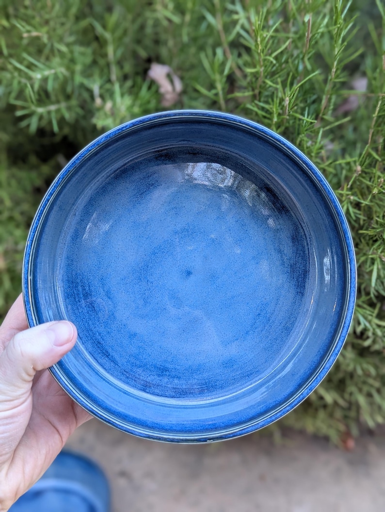May include: A handmade, round ceramic bowl with a deep blue glaze. The bowl has a slightly textured surface and a wide rim with a lighter blue accent. The bowl is held in a hand, with a green leafy background.