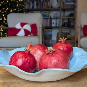 May include: A decorative, light blue ceramic bowl with a wavy rim holds four ripe pomegranates. The pomegranates are a deep red color. The bowl sits on a wooden tray. In the background are two chairs with red and white pillows.