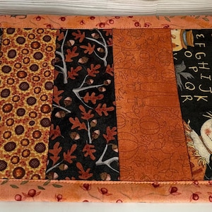 May include: A rectangular, quilted table runner with a patchwork design. The runner features various autumn-themed fabrics, including floral, pumpkin, and leaf patterns in shades of orange, brown, and black. The edges are trimmed with a coordinating fabric and the text "gathering bittersweet".