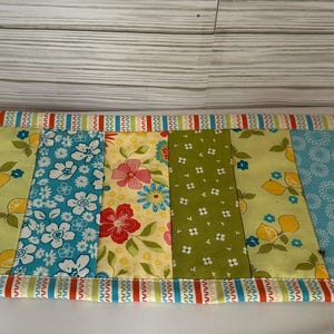 May include: A rectangular quilted table runner with a patchwork design. The runner features vertical panels of various floral and patterned fabrics in blue, yellow, green, and red. The edges are trimmed with a striped border.