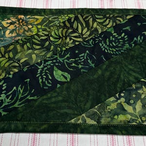May include: A rectangular, quilted placemat with a dark green border and a patchwork design. The patchwork features diagonal stripes of various green fabrics with floral and leaf patterns. The placemat is on a striped surface.